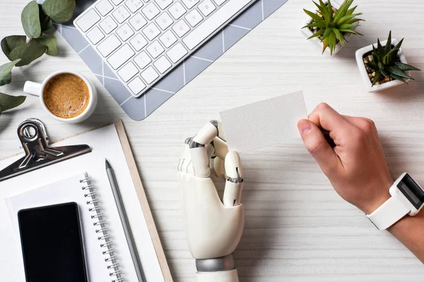 Cropped image of businessman with cyborg hand and smartwatch holding blank visit card at table with smartphone with blank screen in office — Stock Photo