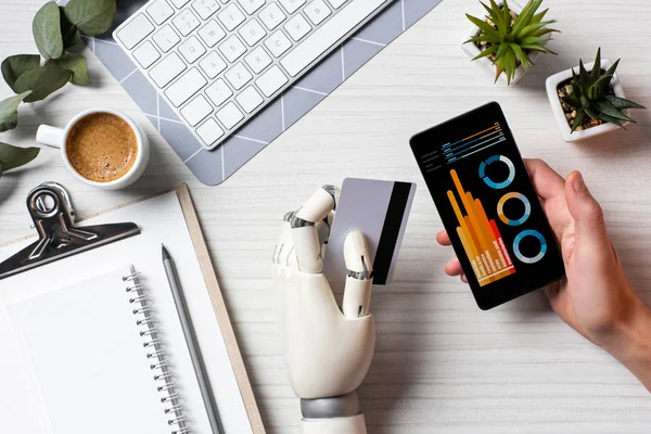 Cropped image of businessman with prosthesis arm holding credit card and using smartphone with infographic on screen at table in office — Stock Photo