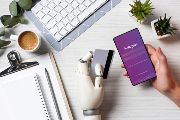 Cropped image of businessman with prosthesis arm holding credit card and using smartphone with instagram on screen at table in office — Stock Photo