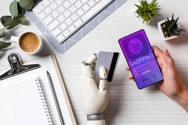 Vista parcial del hombre de negocios con prótesis brazo celebración de la tarjeta de crédito y el uso de teléfono inteligente con compras en la pantalla en la mesa en la oficina - foto de stock