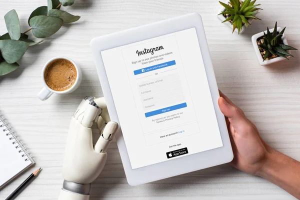 Cropped image of businessman with cyborg hand using digital tablet with instagram on screen at table with coffee cup in office — Stock Photo