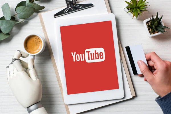 Cropped image of businessman with cyborg hand holding credit card and using digital tablet with youtube on screen at table with coffee cup in office — Stock Photo