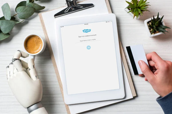 Partial view of businessman with cyborg hand holding credit card and using digital tablet with skype on screen at table with coffee cup in office — Stock Photo