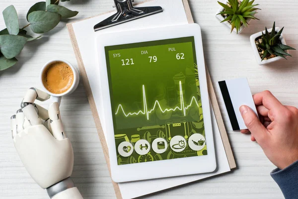 Imagen recortada de hombre de negocios con cyborg mano celebración de la tarjeta de crédito y el uso de la tableta digital con aplicación médica en la pantalla en la mesa con taza de café en la oficina - foto de stock