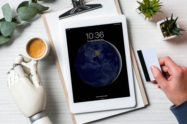 Vista parcial de hombre de negocios con cyborg mano celebración de la tarjeta de crédito y el uso de ipad en la mesa con taza de café en la oficina - foto de stock