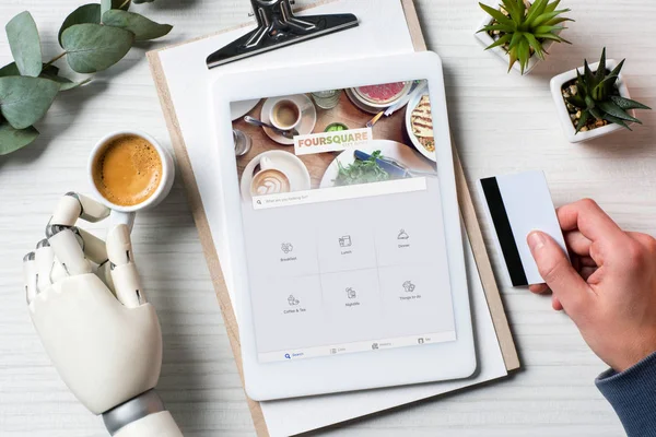 Cropped image of businessman with cyborg hand holding credit card and using digital tablet with foursquare on screen at table with coffee cup in office — Stock Photo