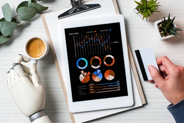 Imagen recortada de hombre de negocios con prótesis mano celebración de tarjeta de crédito y el uso de tableta digital con gráficos en la pantalla en la mesa con taza de café en la oficina - foto de stock