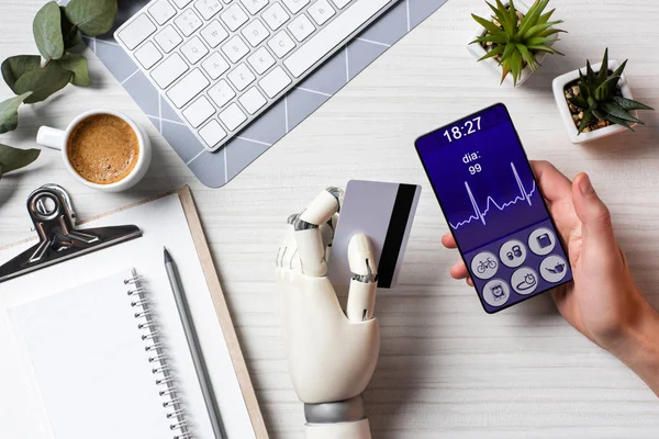 Visão parcial do empresário com mão ciborgue segurando cartão de crédito e usando smartphone com aplicação médica na tela na mesa com xícara de café no escritório — Fotografia de Stock