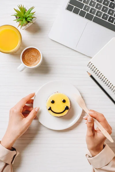 Imagem cortada de empresária comer bolo com símbolo de sorriso à mesa com suco de laranja e xícara de café no escritório — Fotografia de Stock