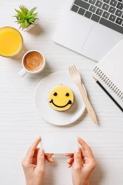 Imagem cortada de empresária segurando cartão de visita vazio à mesa com suco de laranja, xícara de café e bolo com símbolo de sorriso no escritório — Fotografia de Stock