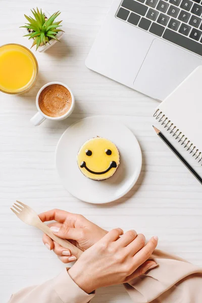 Vista parcial de la mujer de negocios comiendo pastel con símbolo de sonrisa en la mesa con jugo de naranja y taza de café en la oficina - foto de stock