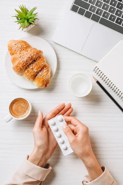 Image recadrée de femme d'affaires prenant des pilules avant le brunch à table avec croissant et tasse de café au bureau — Photo de stock