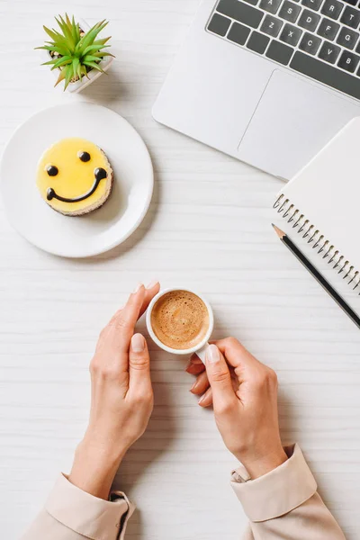 Visão parcial de empresária beber xícara de café à mesa com laptop e bolo com símbolo de sorriso no escritório — Fotografia de Stock