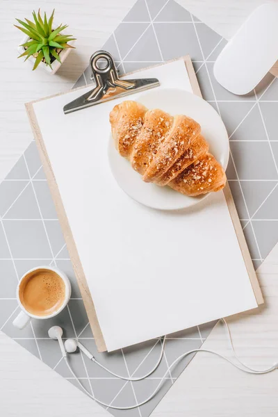 Cama plana con croissant, taza de café, ratón de la computadora y portapapeles en blanco en la mesa en la oficina - foto de stock