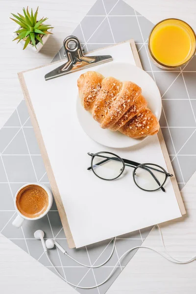 Vue de dessus des lunettes, presse-papiers vide, jus d'orange, tasse à café et croissant à table au bureau — Photo de stock