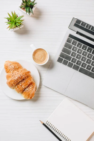 Vista superior del lugar de trabajo con computadora portátil, libro de texto vacío, croissant y taza de café en la oficina - foto de stock