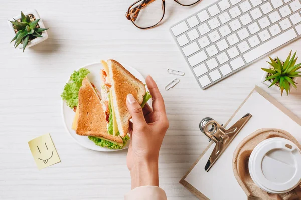 Обрезанный образ деловой женщины, обедающей с бутербродом и кофе в бумажной чашке за столом с символом улыбки в офисе — стоковое фото