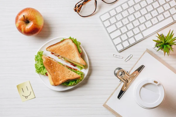 Vista superior del lugar de trabajo con sándwich, taza de café de papel, manzana y símbolo de sonrisa en la mesa en la oficina - foto de stock