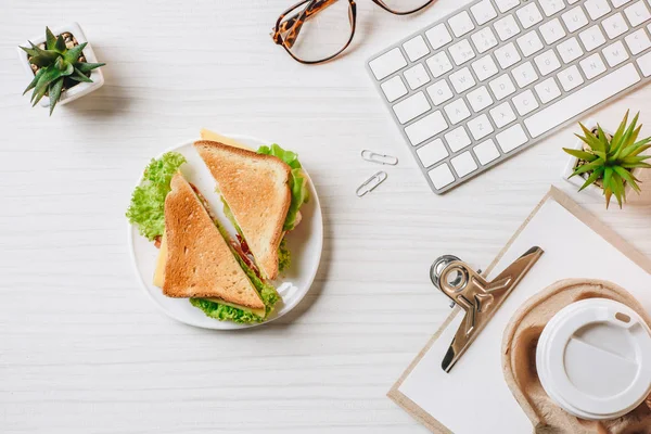 Vue de dessus de tasse à café jetable, sandwich, clavier d'ordinateur et lunettes à la table dans le bureau — Photo de stock