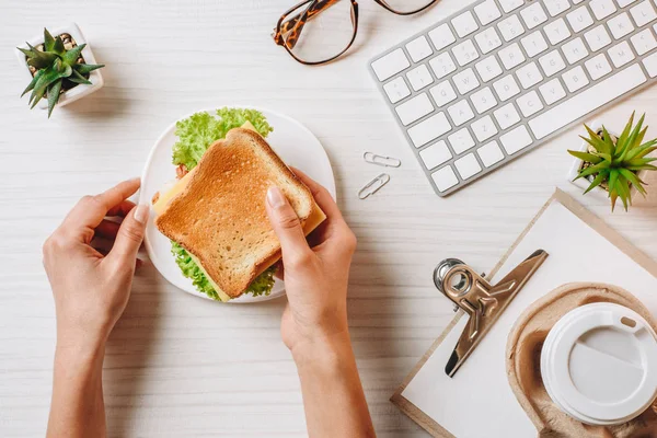 Imagen recortada de la mujer de negocios almorzando con sándwich y café en taza de papel en la mesa con teclado de computadora en la oficina - foto de stock