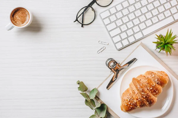 Vista elevada del lugar de trabajo con croissant, taza de café y teclado de la computadora en la oficina - foto de stock