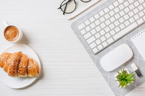 Vue de dessus du clavier d'ordinateur arrangé, des lunettes, de la tasse à café et du croissant à la table au bureau — Photo de stock