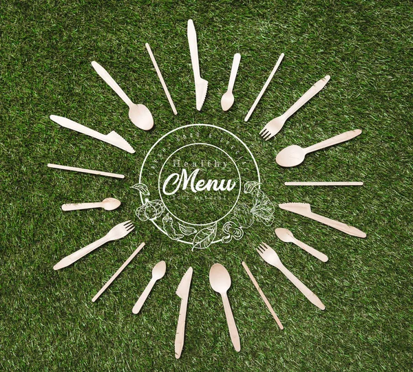 Top view of wooden spoons with forks and knives in shape of sun lying on grass, menu inscription — Stock Photo