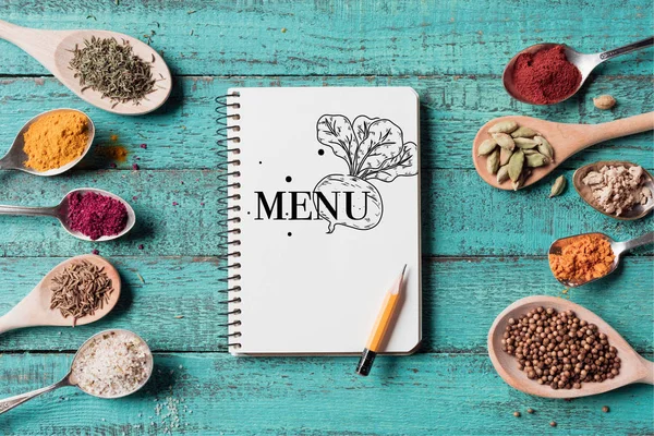 Vista dall'alto del menu iscrizione in notebook con matita e cucchiai con varie spezie su superficie di legno turchese — Foto stock