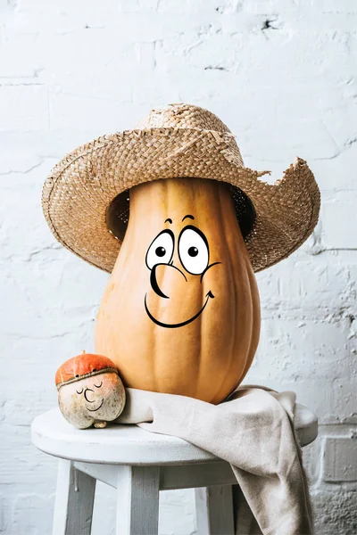 Close up view of ripe pumpkins with drawn happy facial expression and straw hat on wooden stool and white brick wall backdrop — Stock Photo