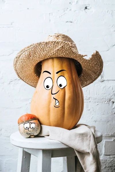 Vista de cerca de calabazas maduras con expresiones faciales dibujadas y sombrero de paja en la superficie de madera y fondo de pared de ladrillo blanco - foto de stock