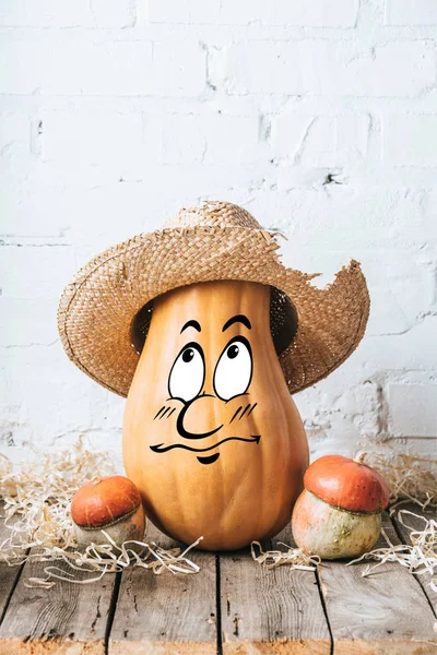 Close up view of ripe pumpkins with drawn bewildered facial expression and straw hat on wooden surface and white brick wall backdrop — Stock Photo