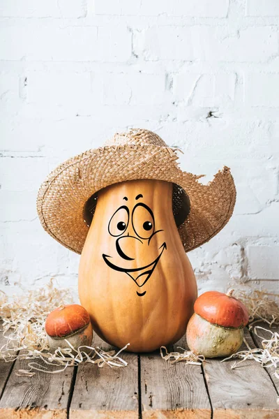 Close up view of ripe pumpkins with drawn smiling facial expression and straw hat on wooden surface and white brick wall backdrop — Stock Photo