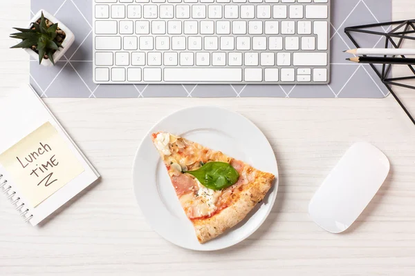 Vista superior de la pizza en el plato, nota con la inscripción de la hora del almuerzo, ratón de la computadora y teclado en el lugar de trabajo - foto de stock