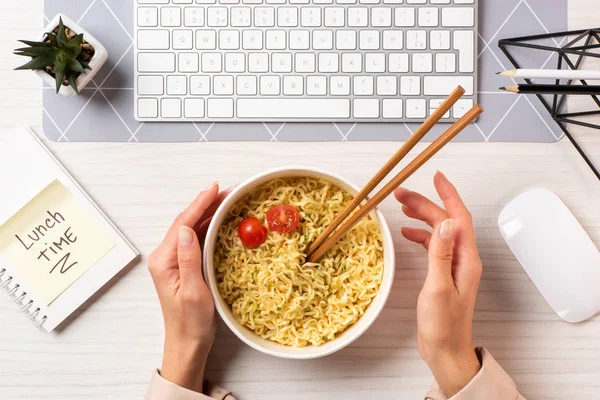 Tiro recortado de la persona sosteniendo tazón con fideos y palillos en el lugar de trabajo - foto de stock