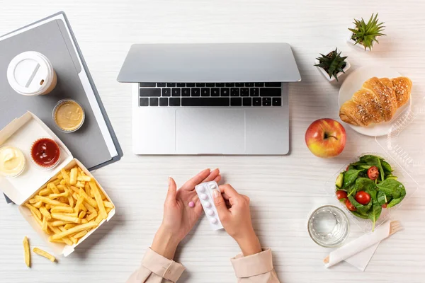 Plan recadré de la personne tenant des pilules sur le lieu de travail avec ordinateur portable, nourriture saine et malbouffe — Photo de stock