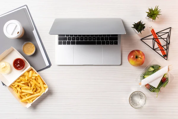 Vista superior de la computadora portátil, comida saludable y comida chatarra en el lugar de trabajo - foto de stock