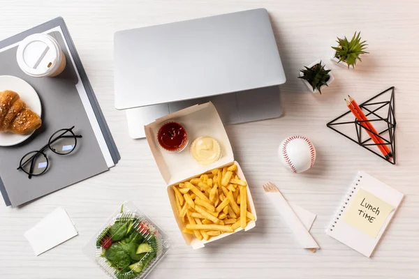 Vista dall'alto di patatine fritte, laptop, insalata di verdure e forniture per ufficio sul posto di lavoro — Stock Photo