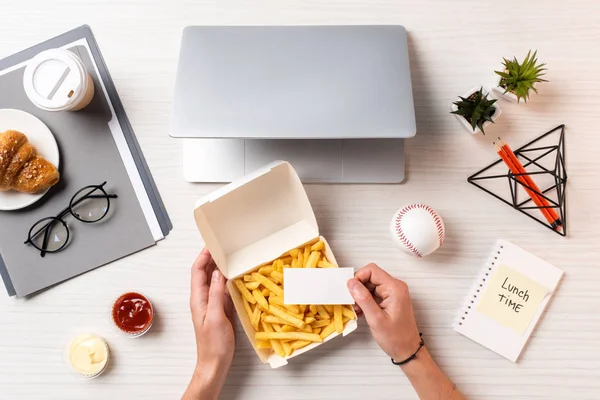 Tiro recortado de la persona que sostiene la tarjeta en blanco encima del envase con papas fritas en el lugar de trabajo - foto de stock