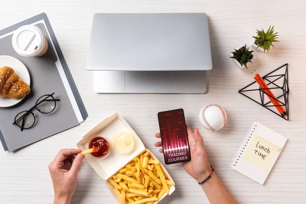 Recortado de la persona comiendo papas fritas con salsa de tomate y el uso de teléfono inteligente con cursos de comercio en el lugar de trabajo - foto de stock