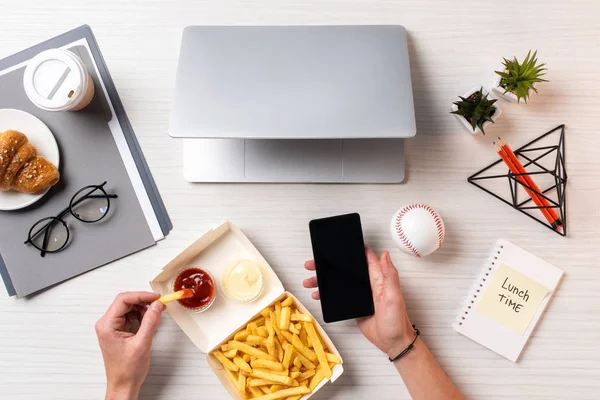 Colpo ritagliato di persona mangiare patatine fritte con ketchup e utilizzando smartphone con schermo bianco sul posto di lavoro — Foto stock