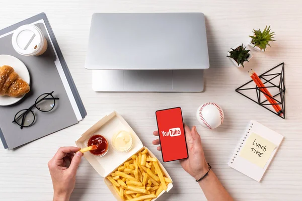 Colpo ritagliato di persona mangiare patatine fritte con ketchup e utilizzando smartphone con app youtube sul posto di lavoro — Foto stock