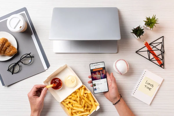 Schnappschuss von Person, die Pommes frites mit Ketchup isst und Smartphone mit Soundcloud-App am Arbeitsplatz nutzt — Stockfoto