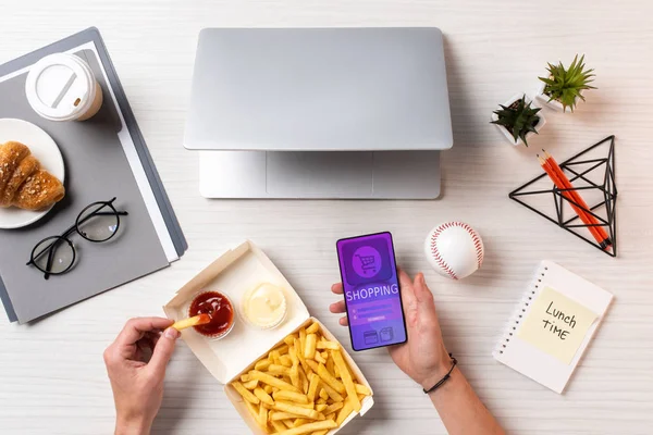 Schnappschuss von Person, die Pommes frites mit Ketchup isst und Smartphone mit Einkaufsanwendung am Arbeitsplatz benutzt — Stockfoto