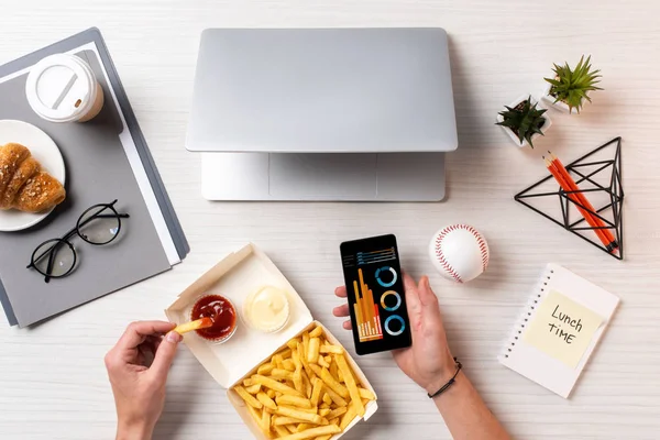 Cropped shot of person eating french fries with ketchup and using smartphone with business charts at workplace — Stock Photo