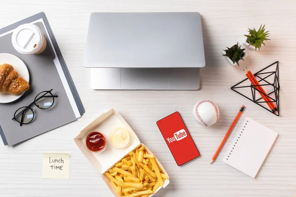 Vista superior de papas fritas, nota adhesiva con la hora del almuerzo inscripción, ordenador portátil y teléfono inteligente con aplicación de youtube en el lugar de trabajo — Stock Photo