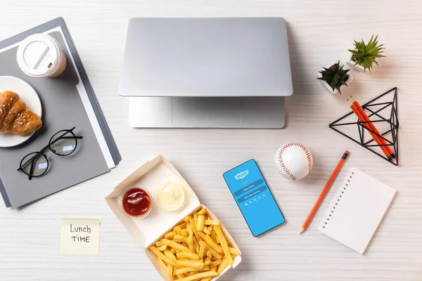 Vista superior de las papas fritas, nota adhesiva con la hora del almuerzo inscripción, ordenador portátil y teléfono inteligente con aplicación de Skype en el lugar de trabajo - foto de stock