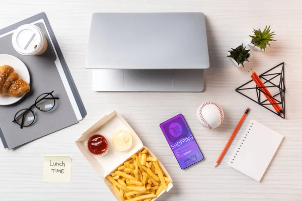 Vista superior de las papas fritas, nota adhesiva con la hora del almuerzo inscripción, ordenador portátil y teléfono inteligente con aplicación de compras en el lugar de trabajo - foto de stock