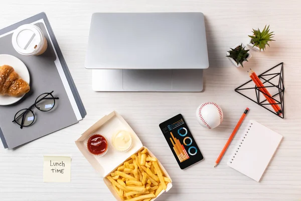Vista superior de batatas fritas, nota pegajosa com inscrição hora do almoço, laptop e smartphone com gráficos no local de trabalho — Fotografia de Stock