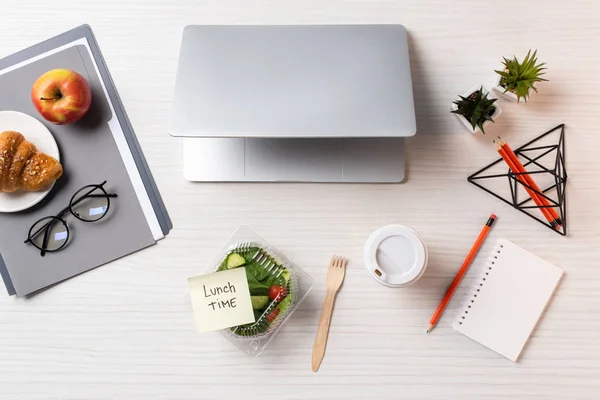 Vista superior do recipiente com salada de legumes e nota pegajosa com inscrição hora do almoço, laptop e material de escritório na mesa — Fotografia de Stock