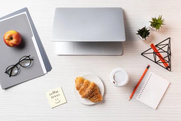 Vista dall'alto di laptop, croissant, tazza di caffè usa e getta e forniture per ufficio sul tavolo — Stock Photo